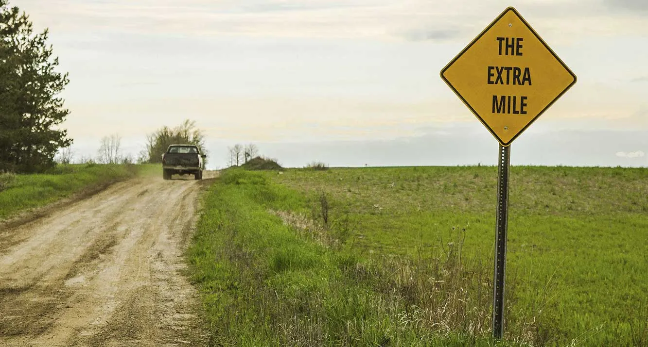 A yellow road sign reading The Extra Mile beside a country road